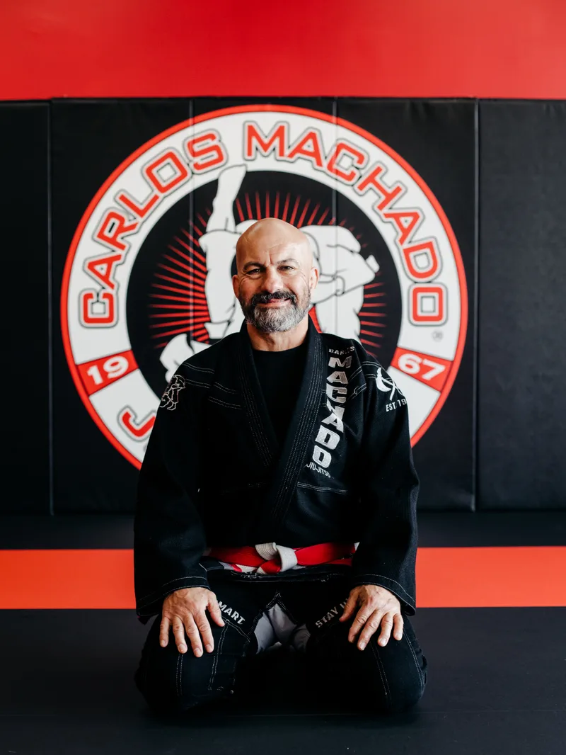 Carlos Machado kneeling in his gi in front of the Carlos Machado academy logo